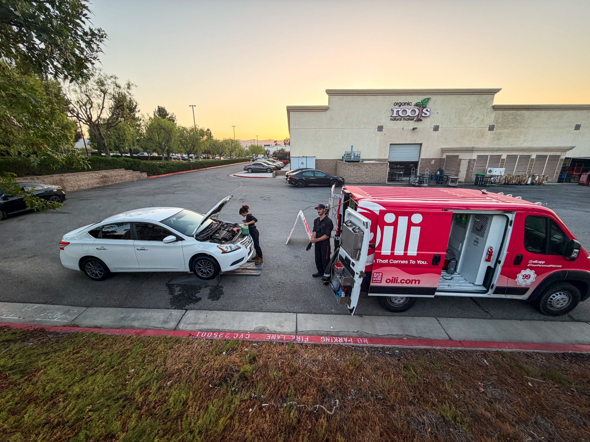 An Oili Pit Crew technician changing a white sedan's oil in a parking lot beside the open Oili van